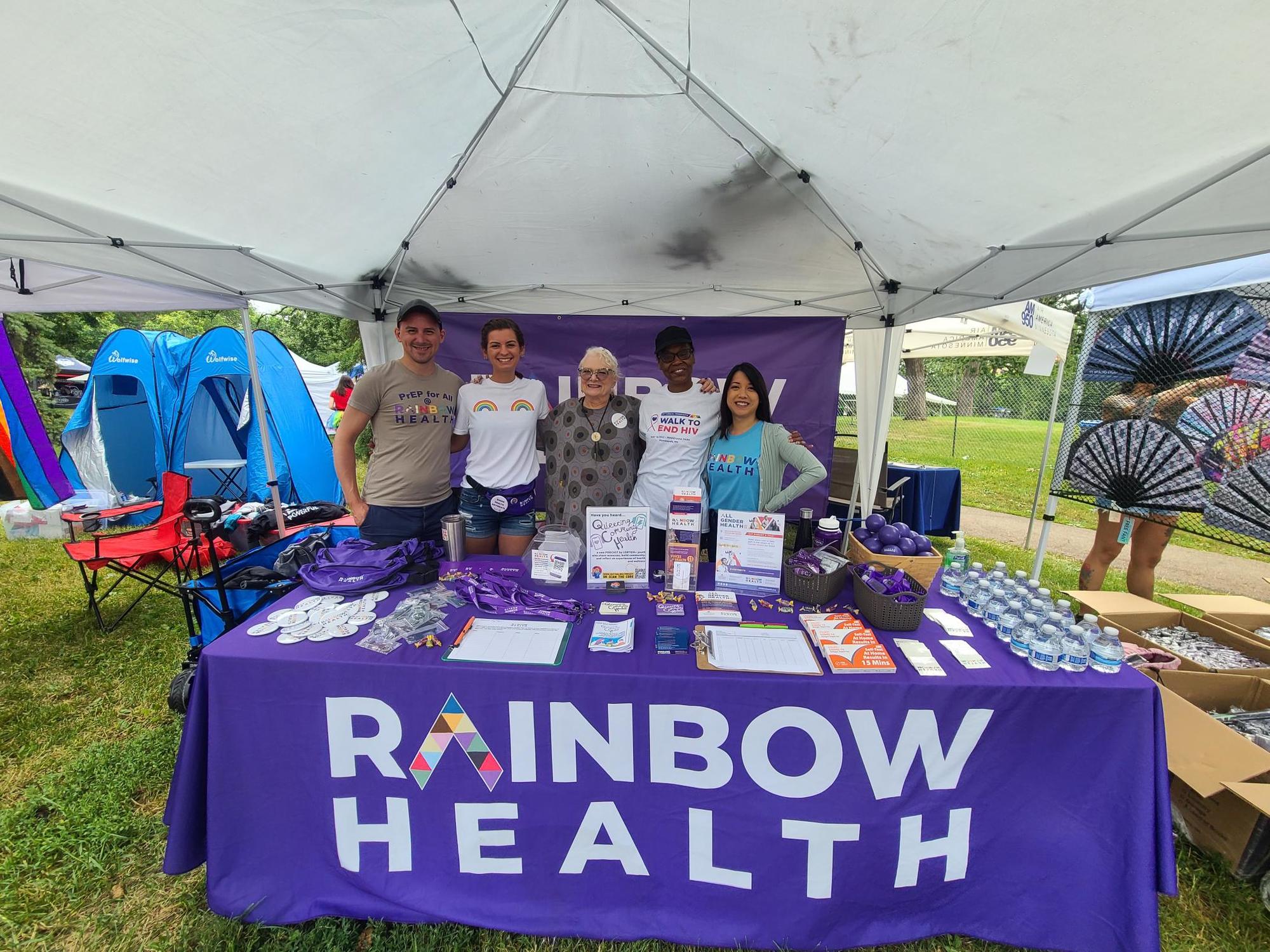 [Picture 1] Rainbow Health staff and volunteers in the booth at the 2022 Twin Cities Pride Festival. Over 500 people visited our booth where they were greeted with an opportunity to win raffle prizes, fun swag items, glitter and skin care kits donated by L’Oreal!